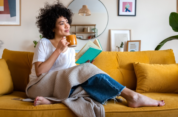 Women reading a book and enjoying a cup of tea on her couch with blanket on her lap.